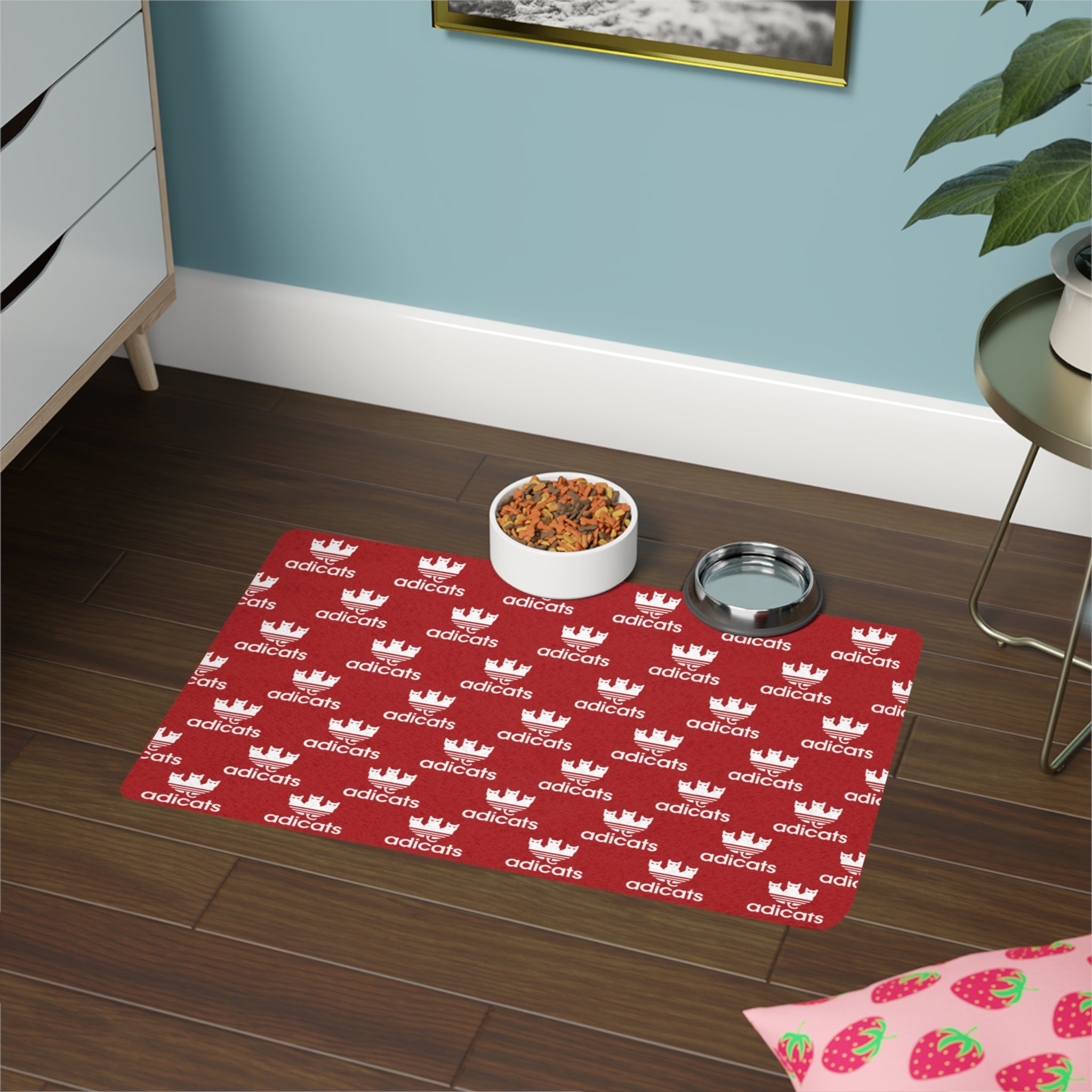 Red cat feeding mat with adicats branding on a wooden floor, featuring a bowl of cat food and a water bowl