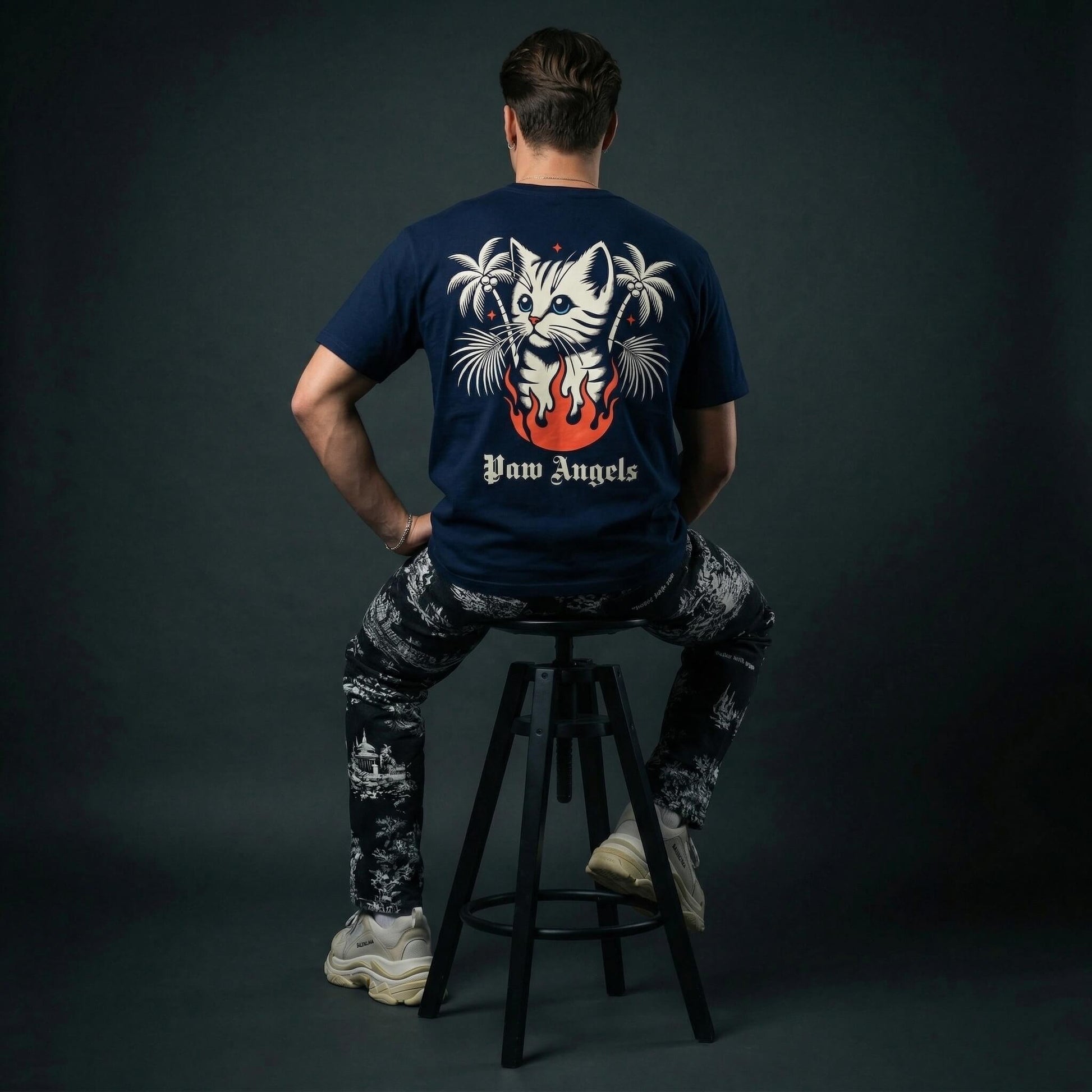 man wearing a navy blue cat themed t-shirt with a graphic design sitting on a stool against a dark background