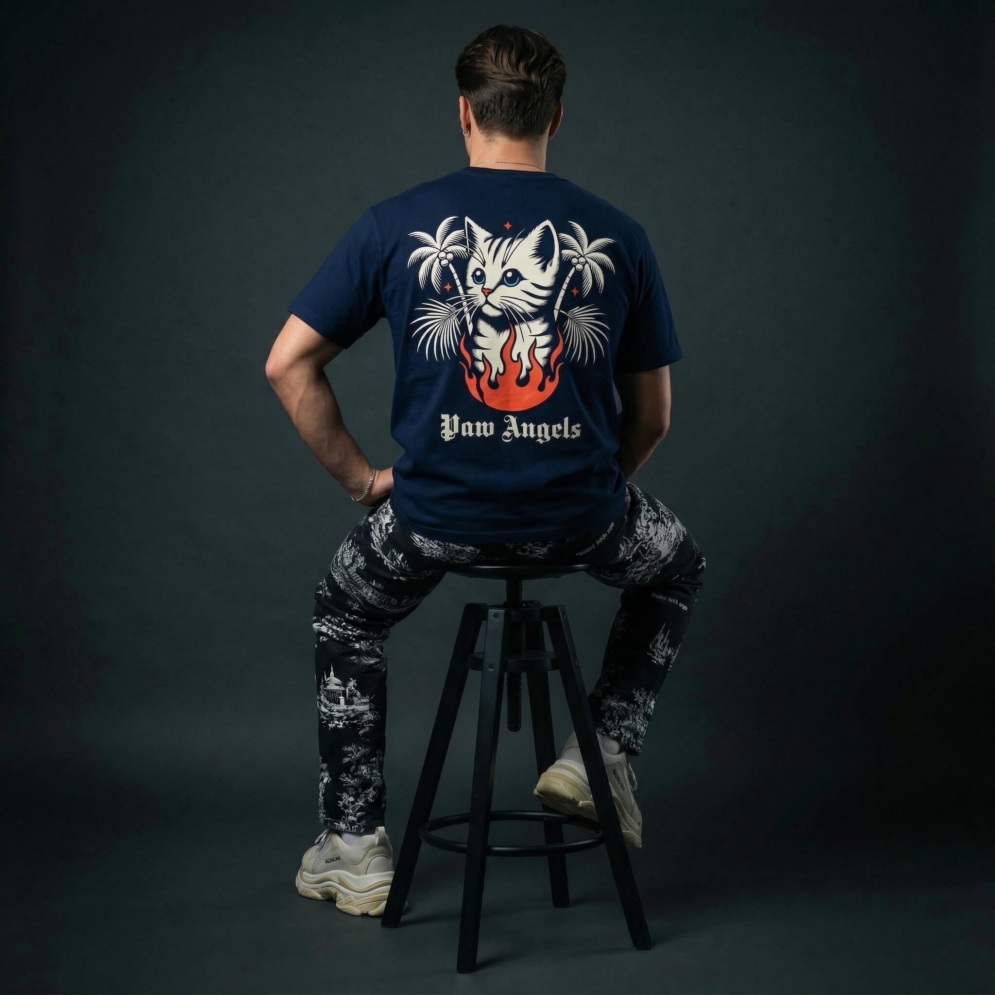 man wearing a navy blue cat themed t-shirt with a graphic design sitting on a stool against a dark background