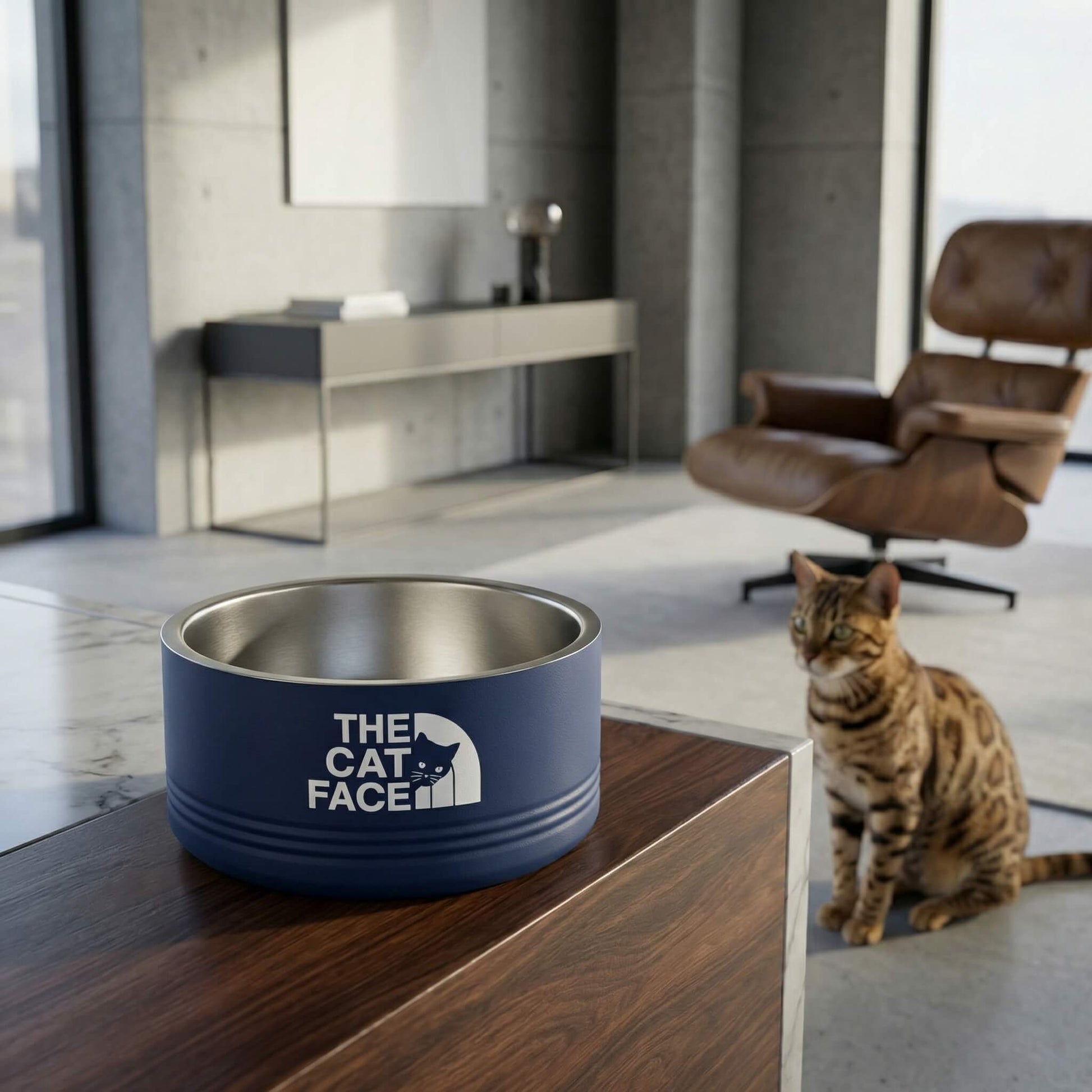 Blue pet bowl with 'The Cat Face' logo on a wooden surface, with a cat sitting in a modern room