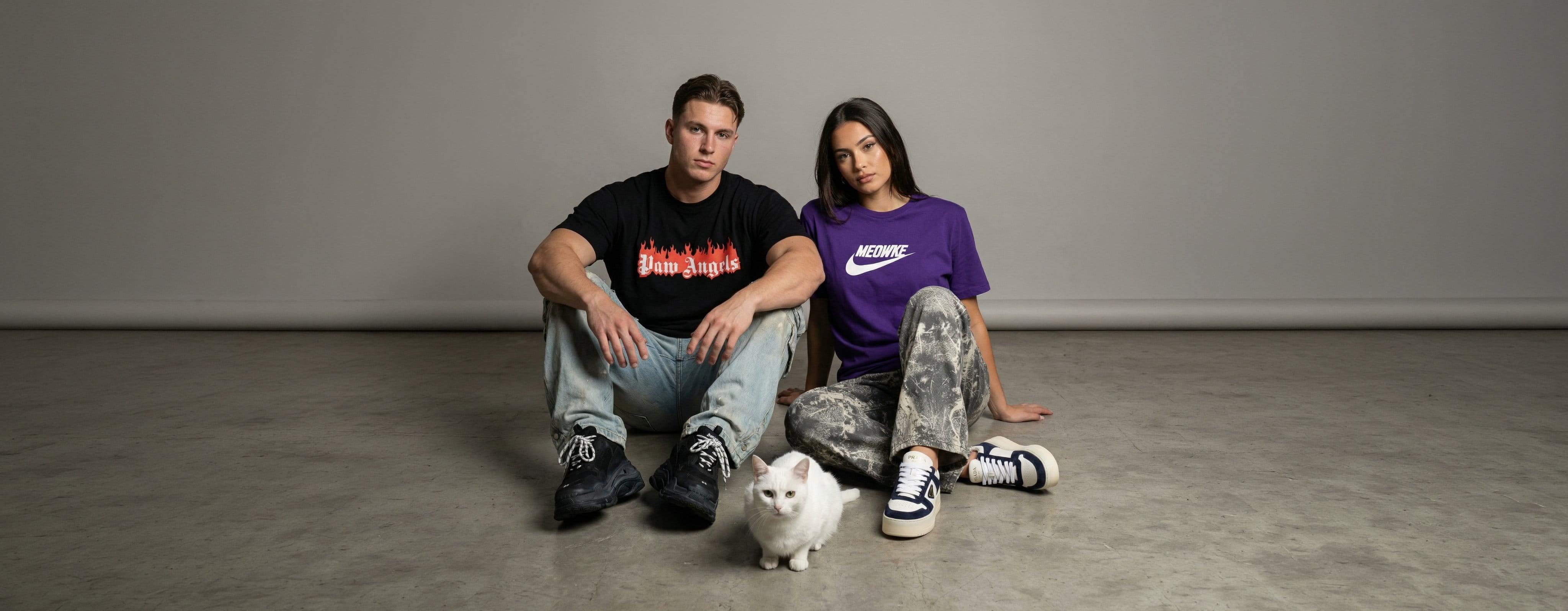 man and woman wearing Mad Cat Cartel funny cat-themed clothing, with a black Paw Angels t shirt and a purple Meowke t shirt beside a white cat, desktop banner image