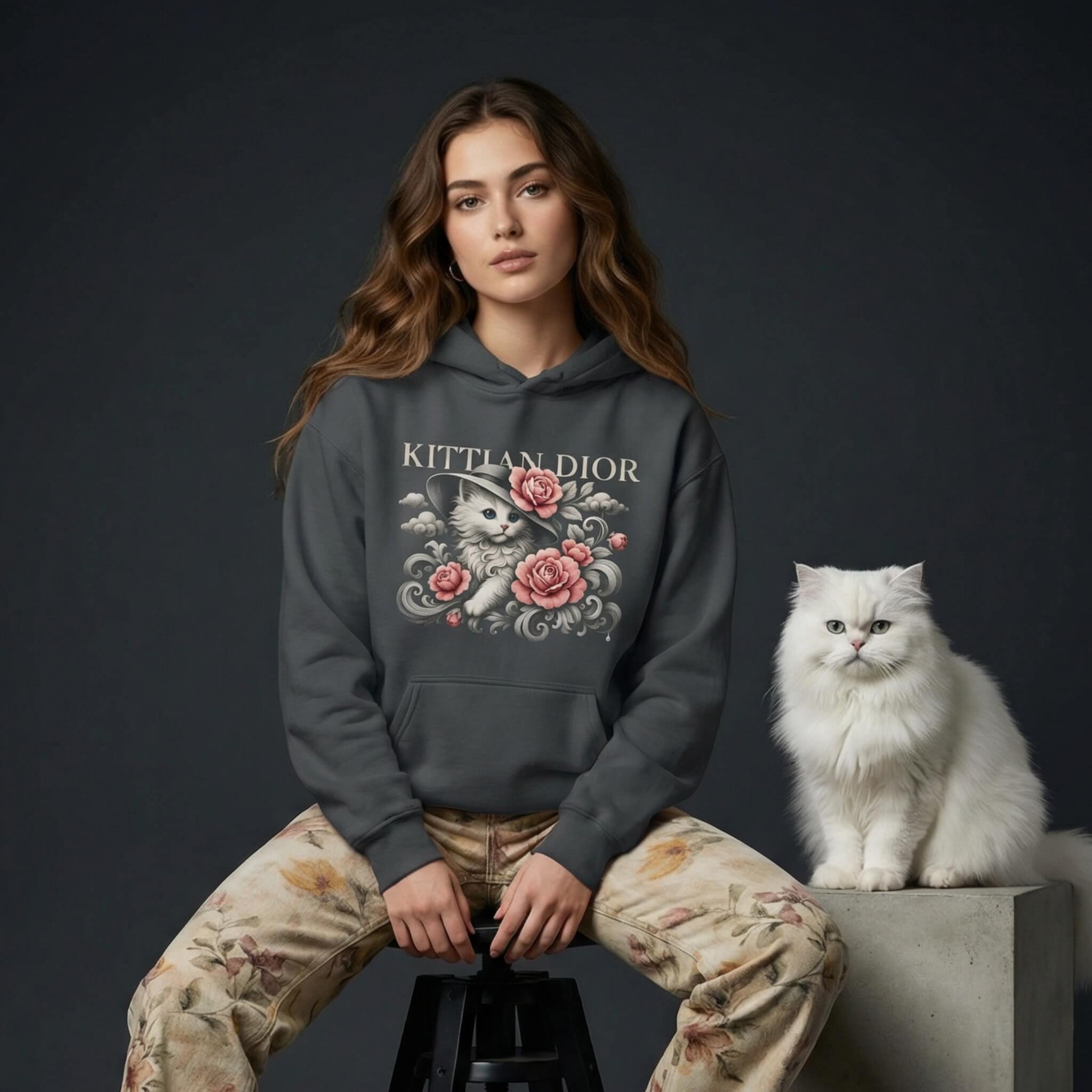 Woman wearing a 'Kittian Dior' hoodie with a cat design, sitting next to a white cat on a dark background